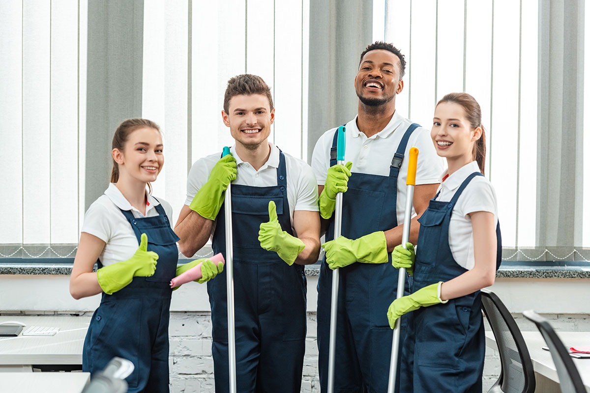 A cheerful team of janitors in uniforms and green gloves posing with cleaning equipment, showcasing their commercial cleaning and janitorial services.