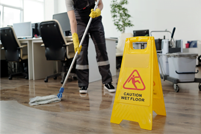 A janitor wearing yellow gloves is mopping the floor in an office with a yellow 'Caution Wet Floor' sign in the foreground, illustrating commercial cleaning and janitorial services.