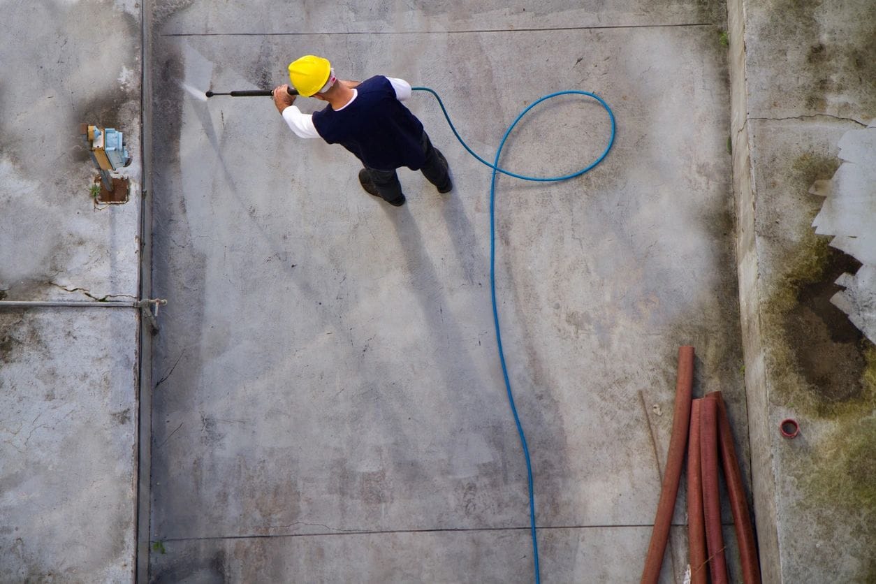 Worker wearing a yellow helmet using a pressure washer to clean a large concrete surface.