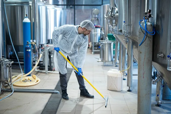 Worker in protective gear cleaning the floor of an industrial facility with a mop, surrounded by stainless steel equipment and storage tanks.