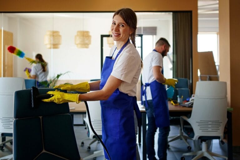 Office cleaning staff in uniform smiling while cleaning chairs in a modern workspace.