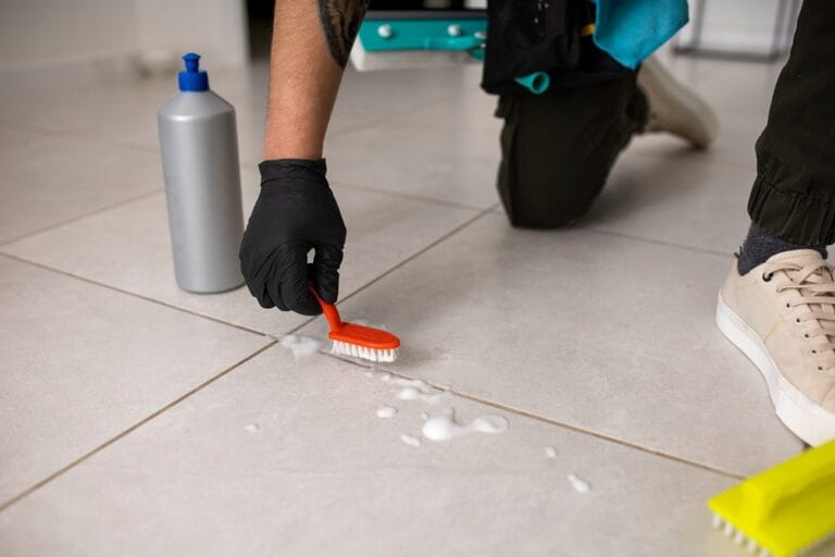 Worker scrubbing grout lines on tile floor with brush and cleaner