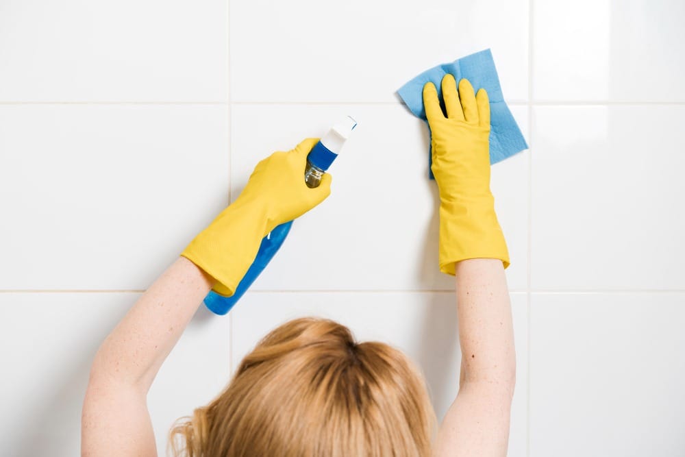Person wearing yellow gloves cleaning tile wall with spray bottle and cloth
