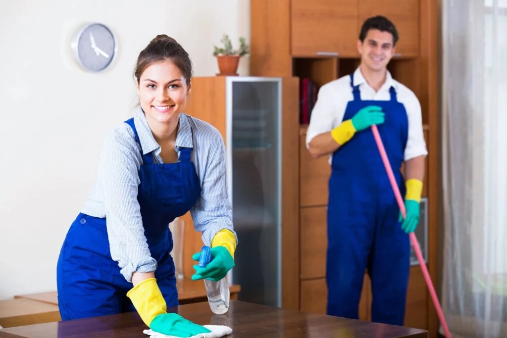 Two professional cleaners in blue uniforms performing custodial and janitorial tasks, including wiping surfaces and mopping floors.